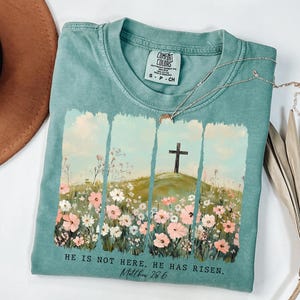 May include: A teal blue t-shirt with a floral design and a cross on a hill. The text "HE IS NOT HERE. HE HAS RISEN. Matthew 28.6" is printed on the bottom of the shirt.