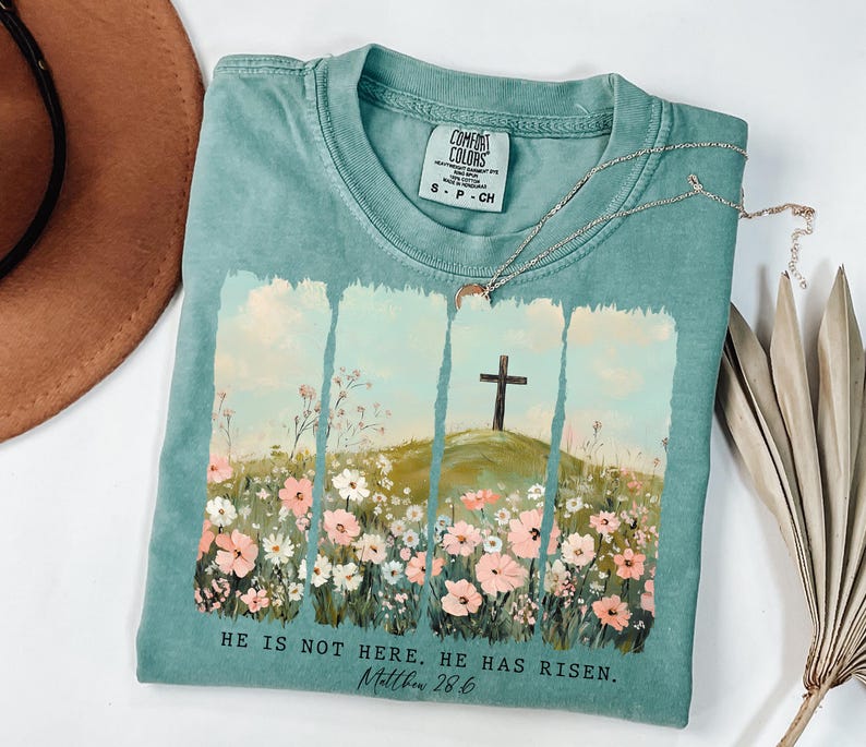 May include: A teal blue t-shirt with a floral design and a cross on a hill. The text "HE IS NOT HERE. HE HAS RISEN. Matthew 28.6" is printed on the bottom of the shirt.