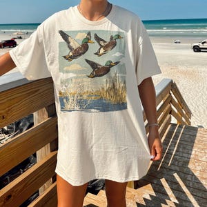 May include: Cream-colored t-shirt featuring a vintage-style graphic of three ducks in flight over a marsh scene. The ducks are brown, green, and white, with a blue and tan background. The shirt has short sleeves.