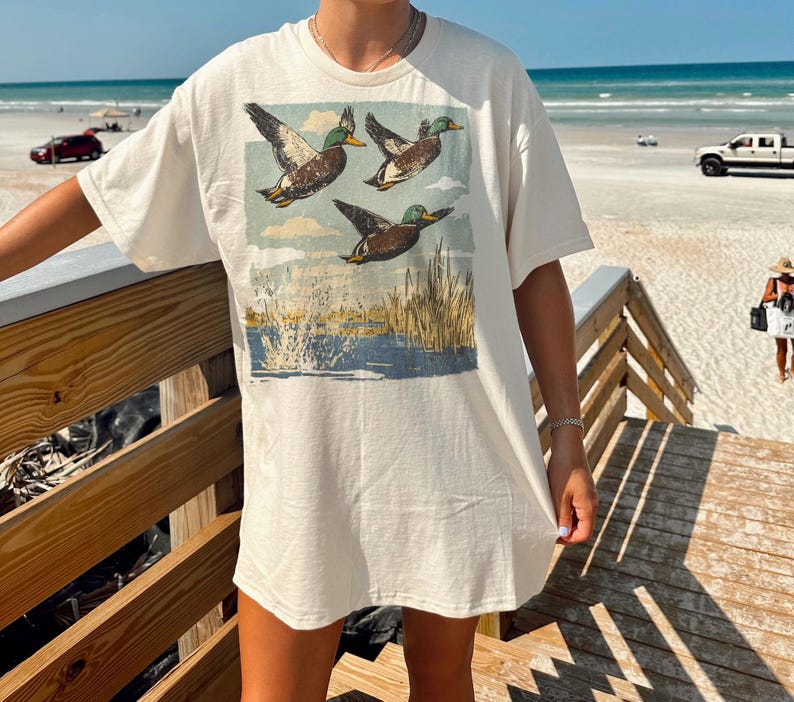 May include: Cream-colored t-shirt featuring a vintage-style graphic of three ducks in flight over a marsh scene. The ducks are brown, green, and white, with a blue and tan background. The shirt has short sleeves.