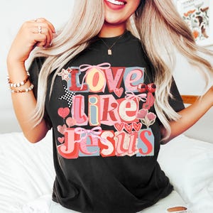 May include: Black t-shirt featuring the phrase "Love Like Jesus" in a colourful, layered design. The graphic incorporates hearts, bows, and floral elements in red, pink, and blue hues.