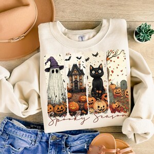 May include: Cream-colored sweatshirt with a Halloween-themed graphic. The design features a ghost wearing a witch hat, a haunted house, a black cat, and jack-o'-lanterns. The words "Spooky Season" are written in a script font.