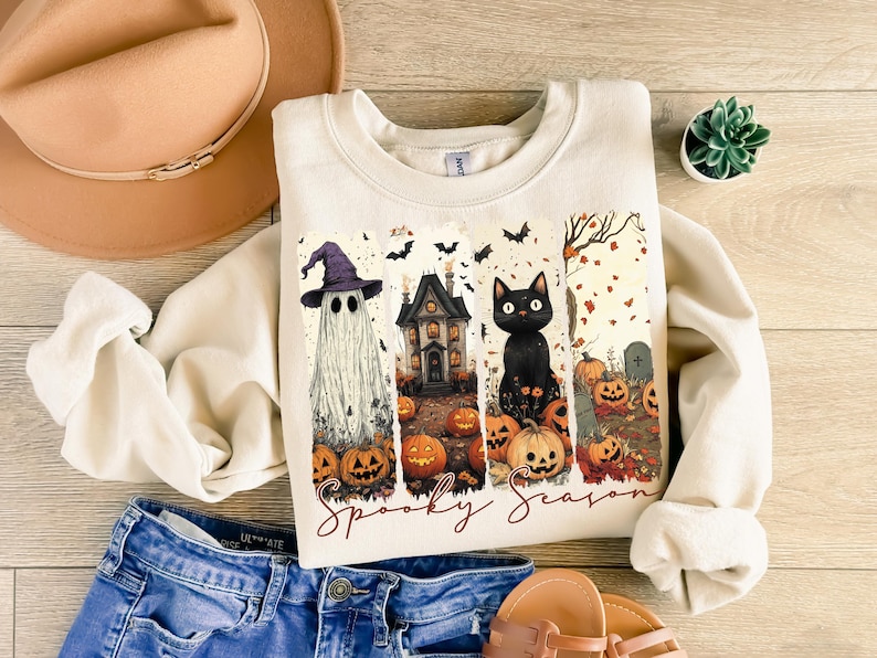 May include: Cream-colored sweatshirt with a Halloween-themed graphic. The design features a ghost wearing a witch hat, a haunted house, a black cat, and jack-o'-lanterns. The words "Spooky Season" are written in a script font.