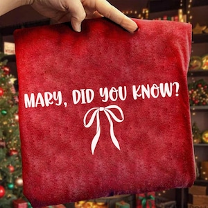 Może przedstawiać: Czerwona torba na prezent o tematyce świątecznej z białym napisem "MARY, DID YOU KNOW?" oraz białą grafiką kokardy. Torba trzymana jest przez dłoń, w tle choinka i dekoracje.