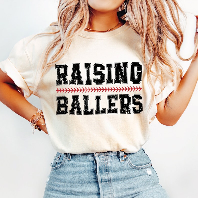May include: A white t-shirt with the text "Raising Ballers" in black with a red baseball stitch design.