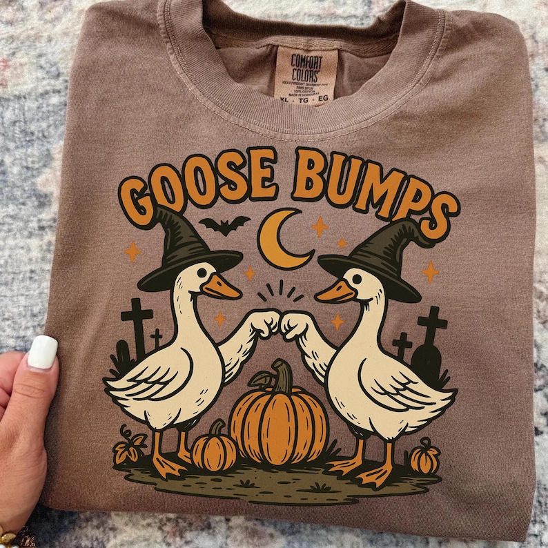 May include: Brown t-shirt with a Halloween-themed graphic. The design features two geese wearing witch hats, bumping fists, with a pumpkin, moon, and the words "GOOSE BUMPS".