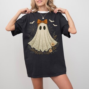 May include: Black t-shirt featuring a ghost design. The ghost is adorned with a brown bow and a lace-like cloak, surrounded by bats, spiderwebs, and floral elements. The design has a vintage, slightly distressed look, perfect for Halloween.
