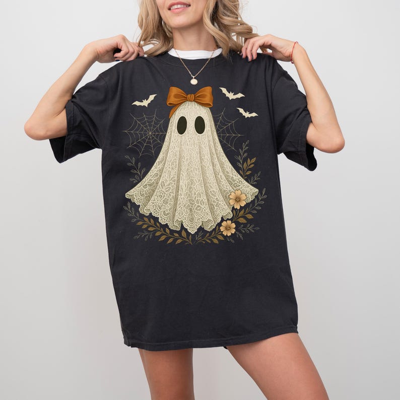 May include: Black t-shirt featuring a ghost design. The ghost is adorned with a brown bow and a lace-like cloak, surrounded by bats, spiderwebs, and floral elements. The design has a vintage, slightly distressed look, perfect for Halloween.