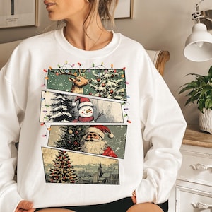 May include: White sweatshirt featuring a Christmas-themed graphic. The design includes panels with a reindeer, snowman, and Santa Claus, along with a Christmas tree and a church. The graphic is framed by a string of colorful lights.