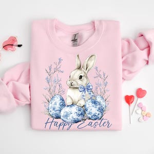 May include: A light pink sweatshirt featuring a watercolor illustration of a white bunny with a blue bow, surrounded by blue patterned Easter eggs and floral accents. The words "Happy Easter" are written in a script font. Heart-shaped sunglasses and lollipops are also visible.