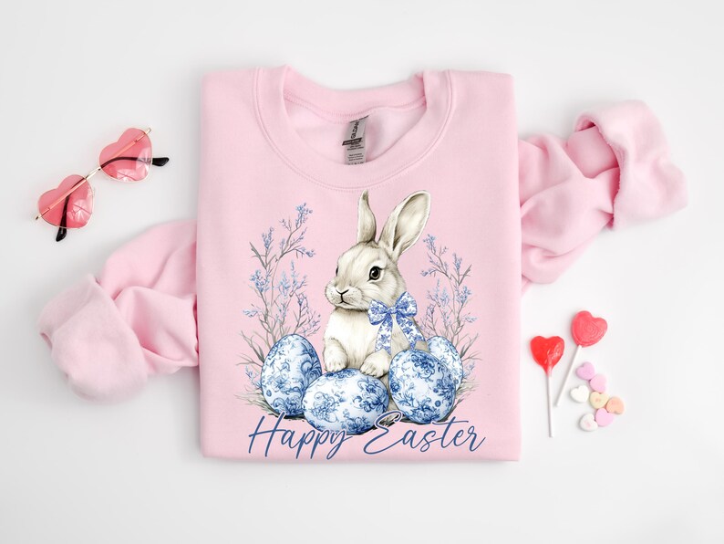 May include: A light pink sweatshirt featuring a watercolor illustration of a white bunny with a blue bow, surrounded by blue patterned Easter eggs and floral accents. The words "Happy Easter" are written in a script font. Heart-shaped sunglasses and lollipops are also visible.