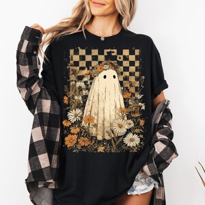 Op de afbeelding: Zwart t-shirt met een spookontwerp, bloemenkrans en bloemen. De achtergrond heeft een dambordpatroon. De persoon draagt een zwart-bruin geruit overhemd.
