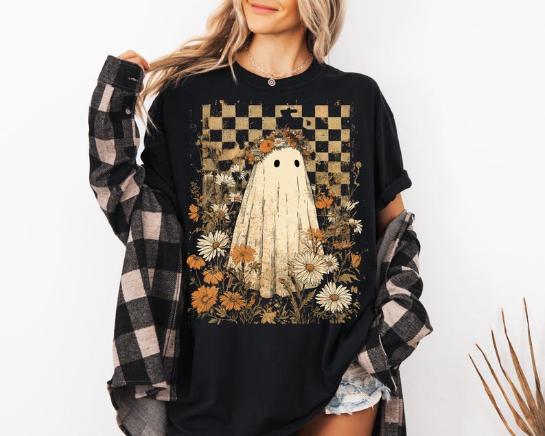 Op de afbeelding: Zwart t-shirt met een spookontwerp, bloemenkrans en bloemen. De achtergrond heeft een dambordpatroon. De persoon draagt een zwart-bruin geruit overhemd.