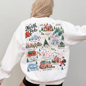 May include: A white long-sleeve sweatshirt featuring a colorful graphic map of the North Pole on the back. The illustration includes labels for Claus Residence, Elf Village, Reindeer Stables, Candy Cane Farm, and Santa's Workshop with Santa.