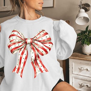 Puede incluir: Sudadera blanca con un gran lazo decorativo. El lazo tiene cintas a rayas rojas y blancas con lunares verdes. La sudadera tiene mangas largas.