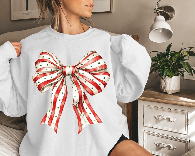 Puede incluir: Sudadera blanca con un gran lazo decorativo. El lazo tiene cintas a rayas rojas y blancas con lunares verdes. La sudadera tiene mangas largas.