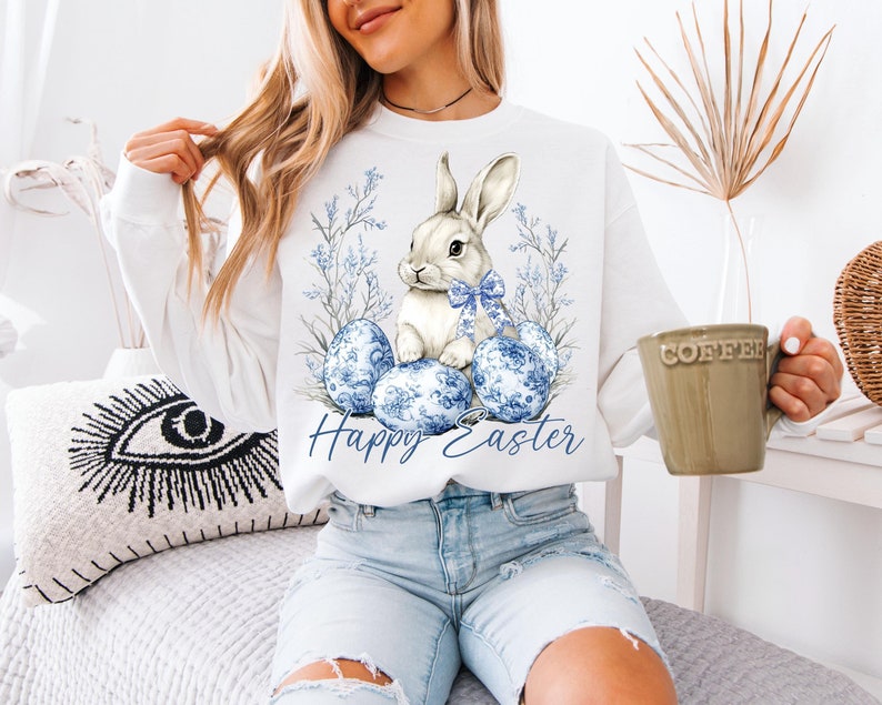 May include: White sweatshirt featuring a watercolor illustration of a bunny with a blue bow, surrounded by blue floral Easter eggs and foliage. The words "Happy Easter" are written in blue script below the design.