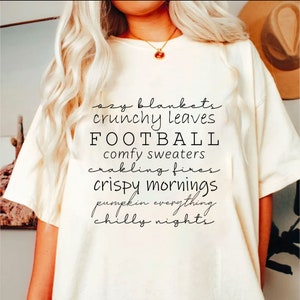 May include: Cream-colored t-shirt with black script text listing fall-themed words: "ozy blankets," "crunchy leaves," "FOOTBALL," "comfy sweaters," "crackling fires," "crispy mornings," "pumpkin everything," and "chilly nights."