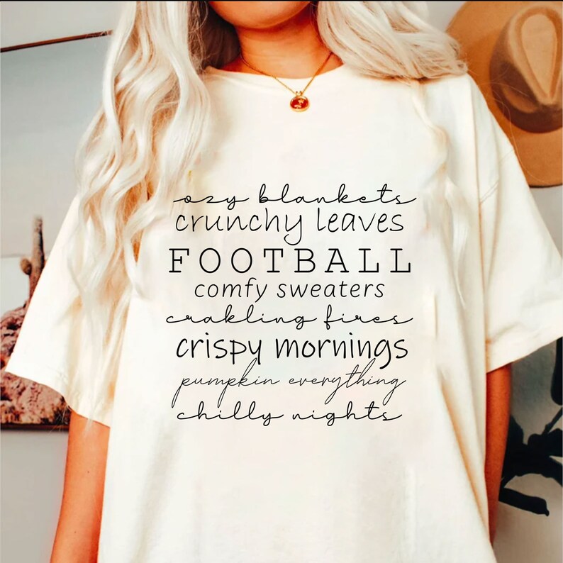 May include: Cream-colored t-shirt with black script text listing fall-themed words: "ozy blankets," "crunchy leaves," "FOOTBALL," "comfy sweaters," "crackling fires," "crispy mornings," "pumpkin everything," and "chilly nights."