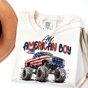 May include: Cream-colored t-shirt featuring a graphic of a monster truck with an American flag design. The text "All American Boy" is printed above the truck in red and blue. A gold necklace and a brown hat are visible.