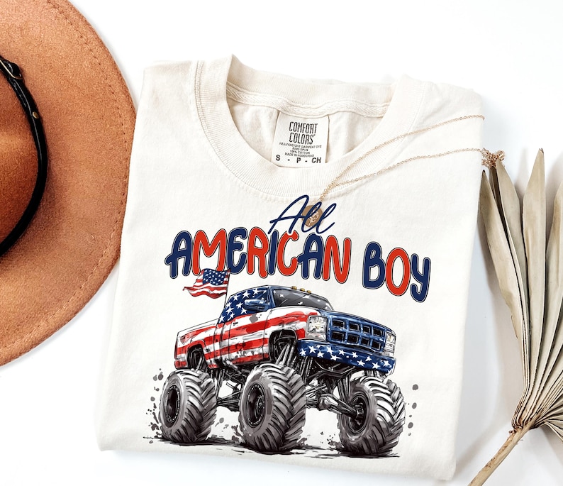May include: Cream-colored t-shirt featuring a graphic of a monster truck with an American flag design. The text "All American Boy" is printed above the truck in red and blue. A gold necklace and a brown hat are visible.
