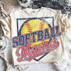 May include: Cream-colored t-shirt featuring a softball graphic. The design includes a yellow softball with red stitching, a blue diamond shape, and the words "SOFTBALL Mama" in blue and red, with a distressed vintage look.