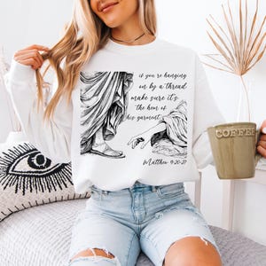 May include: White sweatshirt with a black and white illustration of a hand reaching for the hem of a garment. The text "if you're hanging on by a thread make sure it's the hem of his garment" and "Matthew 9:20-27" are printed below the illustration.