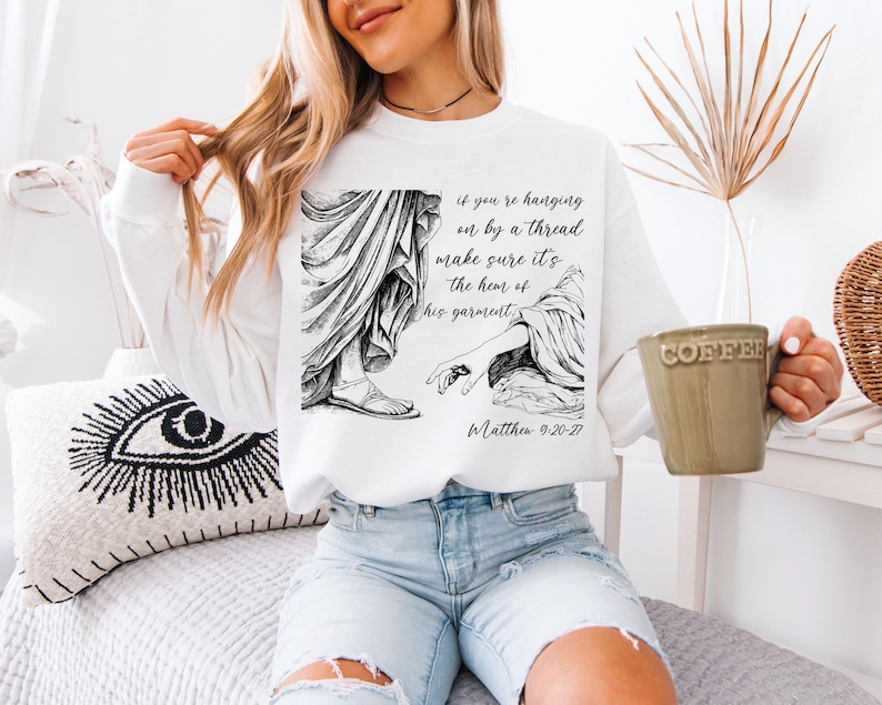 May include: White sweatshirt with a black and white illustration of a hand reaching for the hem of a garment. The text "if you're hanging on by a thread make sure it's the hem of his garment" and "Matthew 9:20-27" are printed below the illustration.
