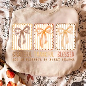 May include: Beige sweatshirt with three rectangular designs featuring bows in shades of brown and orange, set against a striped background. The words "Grateful Thankful Blessed" are printed below the designs, with the text "God is faithful in every season."