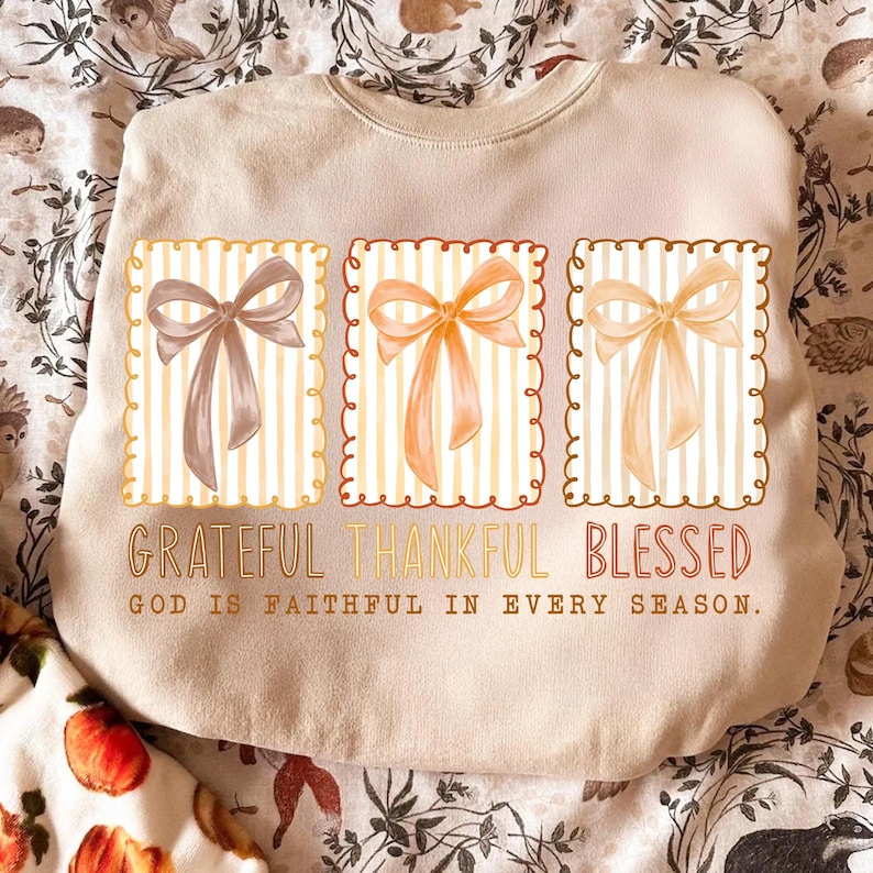 May include: Beige sweatshirt with three rectangular designs featuring bows in shades of brown and orange, set against a striped background. The words "Grateful Thankful Blessed" are printed below the designs, with the text "God is faithful in every season."
