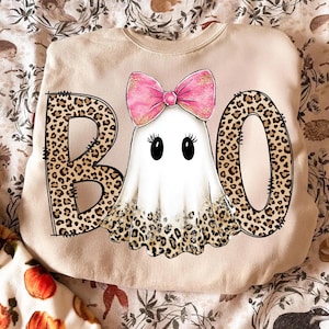 May include: Beige sweatshirt with a Halloween-themed design. The word "BOO" is spelled out with leopard print letters. A cartoon ghost with a pink bow is in the center. The ghost has black eyes and a leopard print bottom.