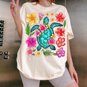Pu&ograve; includere: Una maglietta bianca con una grafica colorata di una tartaruga marina circondata da fiori tropicali. La tartaruga &egrave; blu turchese e verde, e i fiori sono rosa, rossi, gialli e viola.