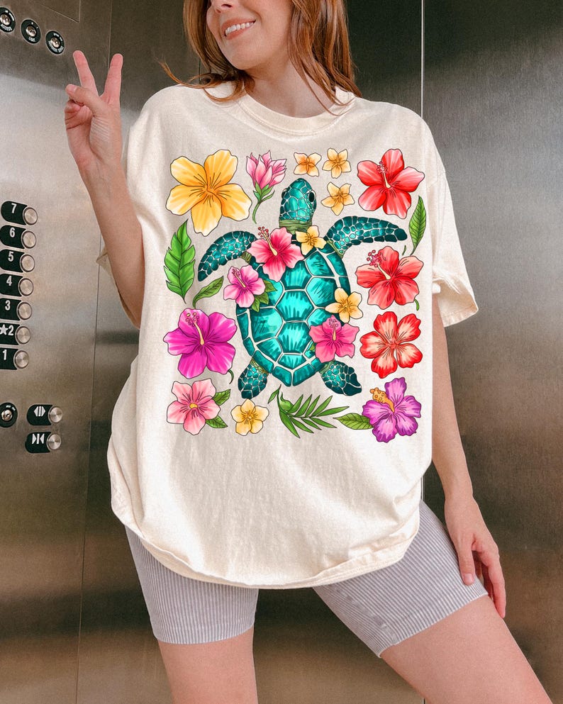 Pu&ograve; includere: Una maglietta bianca con una grafica colorata di una tartaruga marina circondata da fiori tropicali. La tartaruga &egrave; blu turchese e verde, e i fiori sono rosa, rossi, gialli e viola.