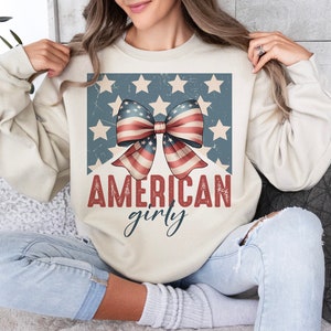 May include: A cream-colored sweatshirt with a distressed graphic of a red, white, and blue bow with stars on a blue background. The text "AMERICAN girly" is printed below the bow.
