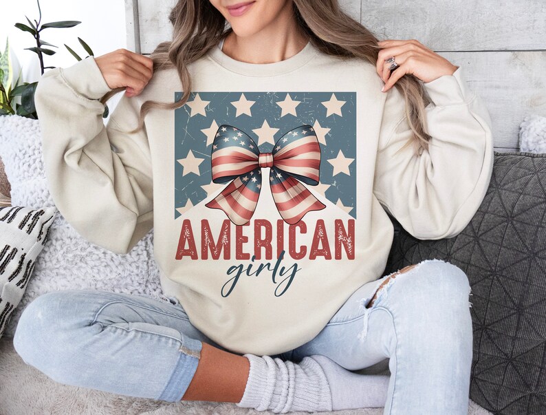 May include: A cream-colored sweatshirt with a distressed graphic of a red, white, and blue bow with stars on a blue background. The text "AMERICAN girly" is printed below the bow.