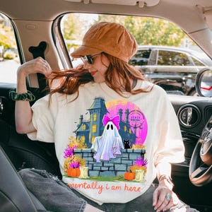 Puede incluir: Una camiseta color crema con un gr&aacute;fico de un fantasma con un lazo rosa sentado en un muro de piedra frente a una casa encantada. El dise&ntilde;o incluye un gato negro, calabazas naranjas y el texto "mentally i am here" en letra naranja.