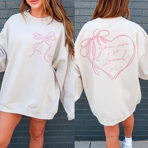 May include: A beige sweatshirt with a pink ribbon bow design on the front and a pink heart with the text "you are so loved John 3:16" on the back.