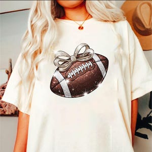 Pode incluir: Camiseta branca com uma bola de futebol marrom e branca com glitter e um la&ccedil;o prateado no topo.