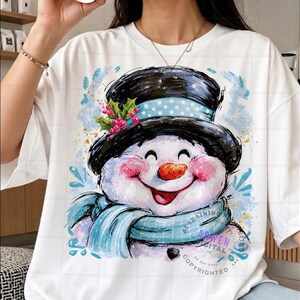 May include: A white t-shirt featuring a graphic illustration of a smiling snowman wearing a black top hat with a light blue polka dot band, a light blue scarf, and a sprig of holly with red berries, accented by blue paint-like splashes.