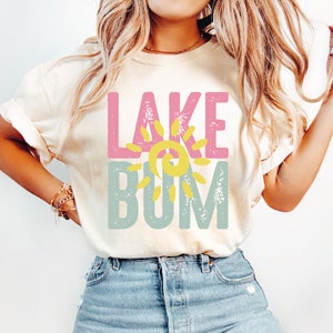 Op de afbeelding: Een wit T-shirt met de tekst "LAKE" in roze en "BUM" in blauw met een gele zon-afbeelding ertussen.
