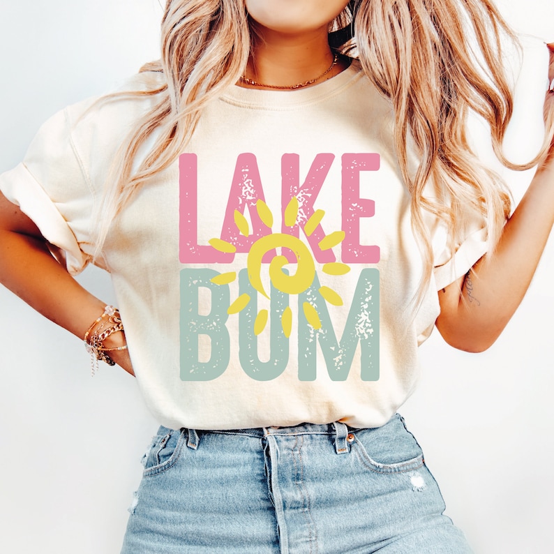 Op de afbeelding: Een wit T-shirt met de tekst "LAKE" in roze en "BUM" in blauw met een gele zon-afbeelding ertussen.