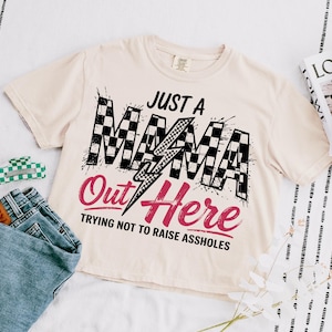 Puede incluir: Una camiseta beige con un gr&aacute;fico a cuadros en blanco y negro que dice "Just a Mama Out Here Trying Not to Raise Assholes".