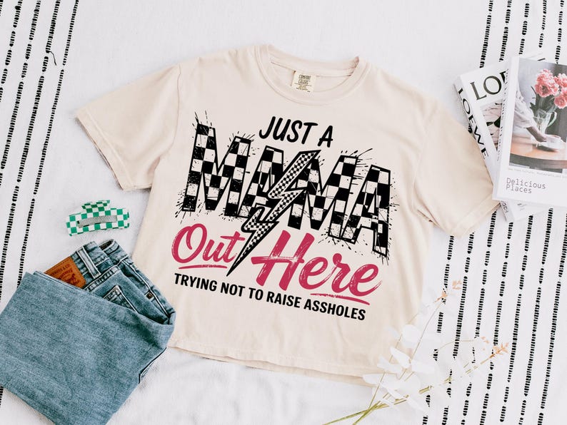 Puede incluir: Una camiseta beige con un gr&aacute;fico a cuadros en blanco y negro que dice "Just a Mama Out Here Trying Not to Raise Assholes".