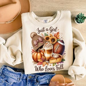 K&ouml;nnte beinhalten: Cremefarbenes Sweatshirt mit dem Text "Just a Girl Who Loves Fall" und einer Grafik mit Herbstmotiven. Die Grafik zeigt einen K&uuml;rbis, einen Football, einen Latte, ein St&uuml;ck Kuchen und eine M&uuml;tze.