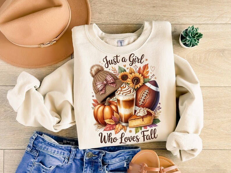 K&ouml;nnte beinhalten: Cremefarbenes Sweatshirt mit dem Text "Just a Girl Who Loves Fall" und einer Grafik mit Herbstmotiven. Die Grafik zeigt einen K&uuml;rbis, einen Football, einen Latte, ein St&uuml;ck Kuchen und eine M&uuml;tze.