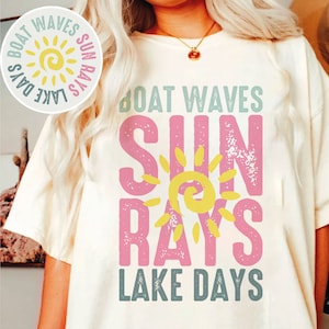Op de afbeelding: Een wit T-shirt met een roze en geel grafisch ontwerp met de tekst "Boat Waves Sun Rays Lake Days". De grafiek toont een gestileerde zon met een gele swirl.