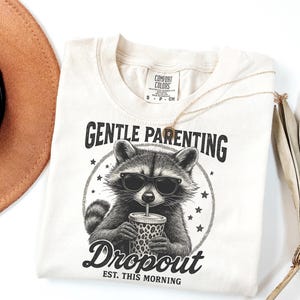 Puede incluir: Camiseta color crema con un gr&aacute;fico de un mapache con gafas de sol y una taza con estampado de leopardo. El texto dice "GENTLE PARENTING" encima del mapache y "Dropout EST. THIS MORNING" debajo.