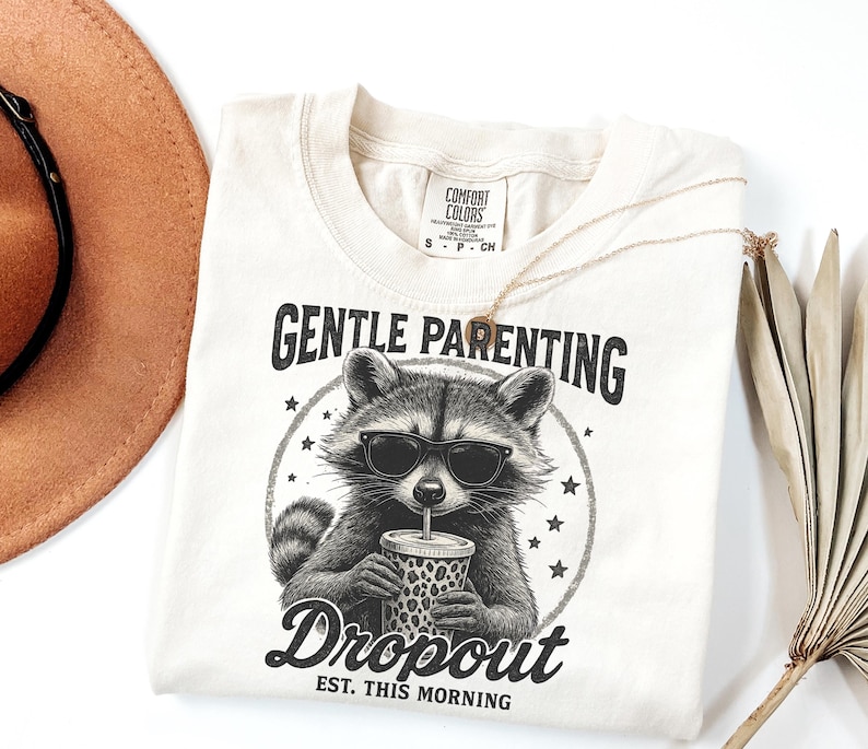 Puede incluir: Camiseta color crema con un gr&aacute;fico de un mapache con gafas de sol y una taza con estampado de leopardo. El texto dice "GENTLE PARENTING" encima del mapache y "Dropout EST. THIS MORNING" debajo.
