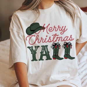 May include: Cream-colored t-shirt with a Christmas-themed graphic. The design features a green cowboy hat, the text "Merry Christmas Y'all" in red and green, and green cowboy boots decorated with candy canes.