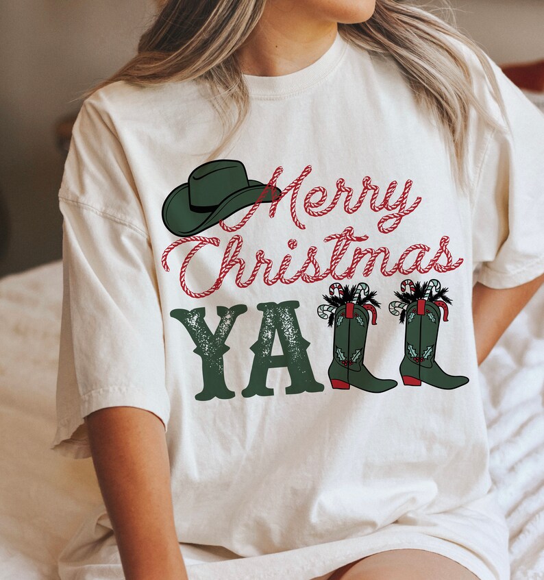 May include: Cream-colored t-shirt with a Christmas-themed graphic. The design features a green cowboy hat, the text "Merry Christmas Y'all" in red and green, and green cowboy boots decorated with candy canes.